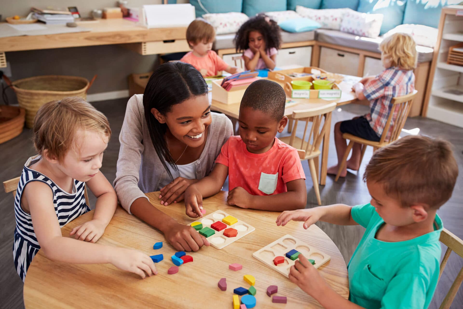 A teacher guiding four children through a shape-sorting activity at a classroom table