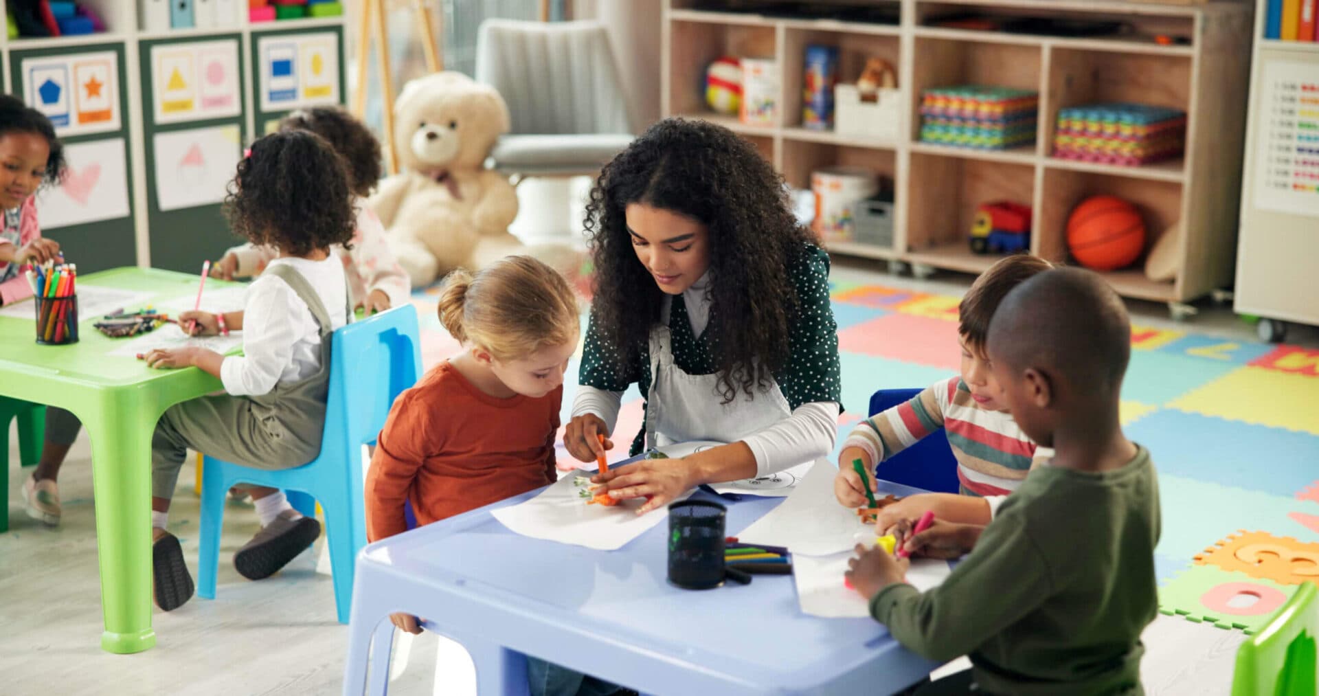 A teacher kneeling at a low table helping two children with a cutting and gluing activity