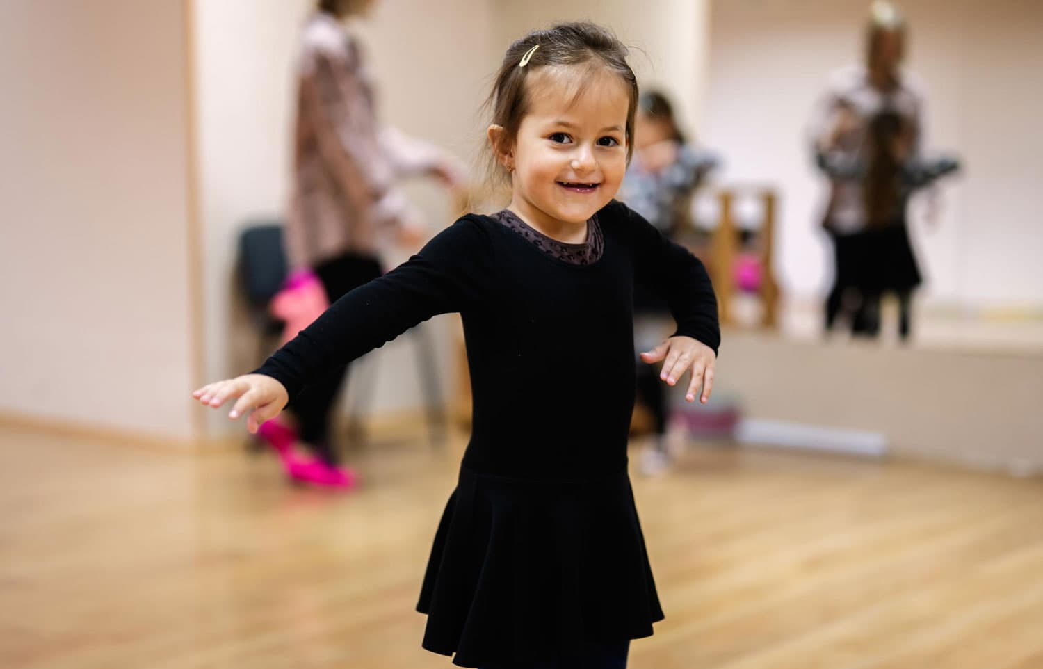A young girl in a black leotard dancing in a mirrored studio with classmates behind her
