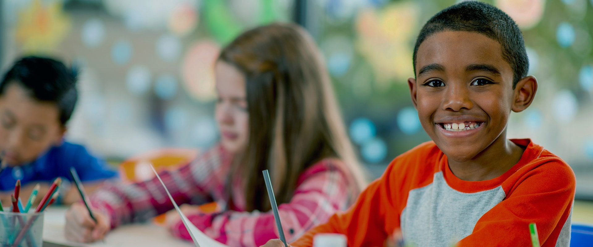 Young child smiling at classroom desk with paper and pencil.