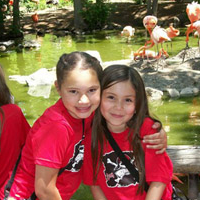Two children sitting outside by a pond smiling with arms around each other.